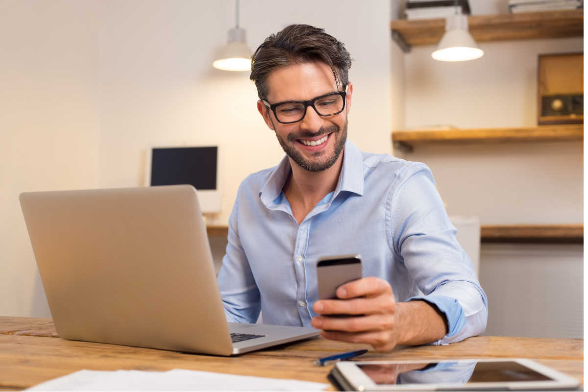 A smiling man sitting in front of his laptop and looking at his phone